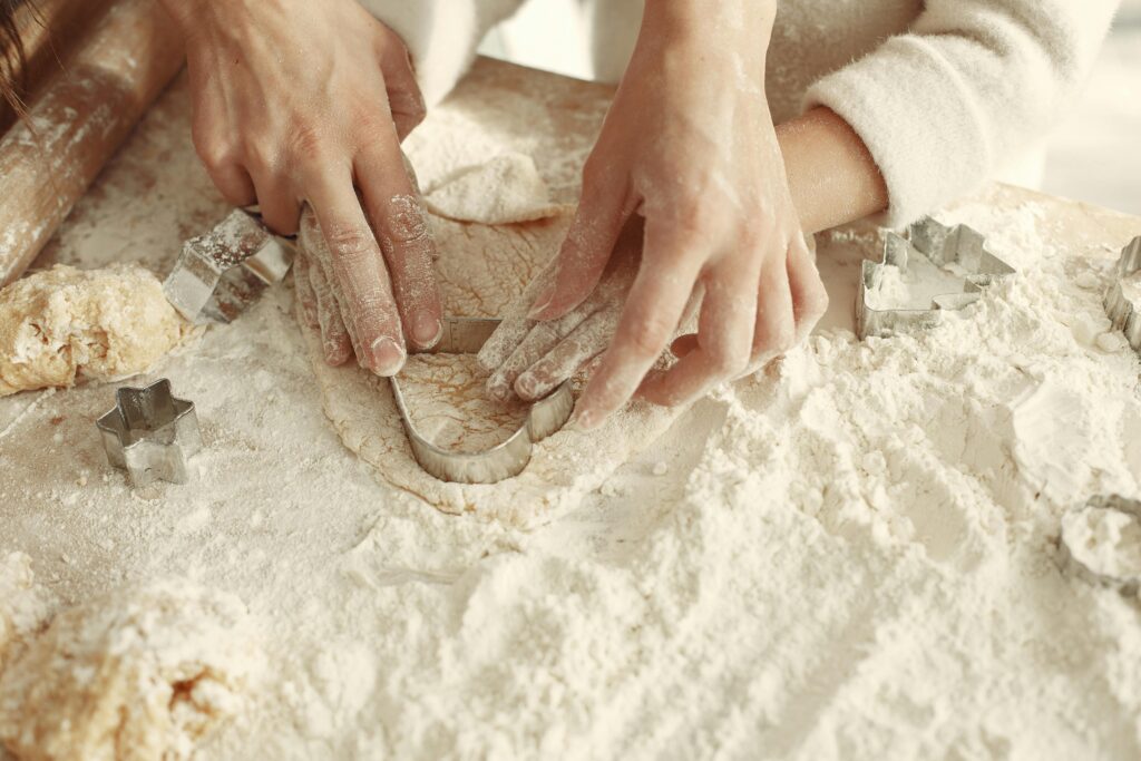 Hands of a parent and child using cookie cutters on dough during a baking session.